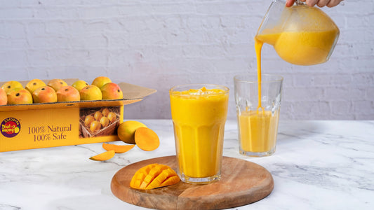 A glass of fresh mango smoothie, with a box of mangoes and sliced fruit on a marble tabletop.