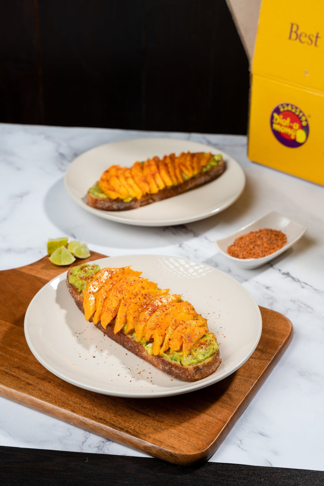 Avocado toast topped with sliced Alphonso mango, chilli seasoning, and lime wedges on a plate with a side of spices.