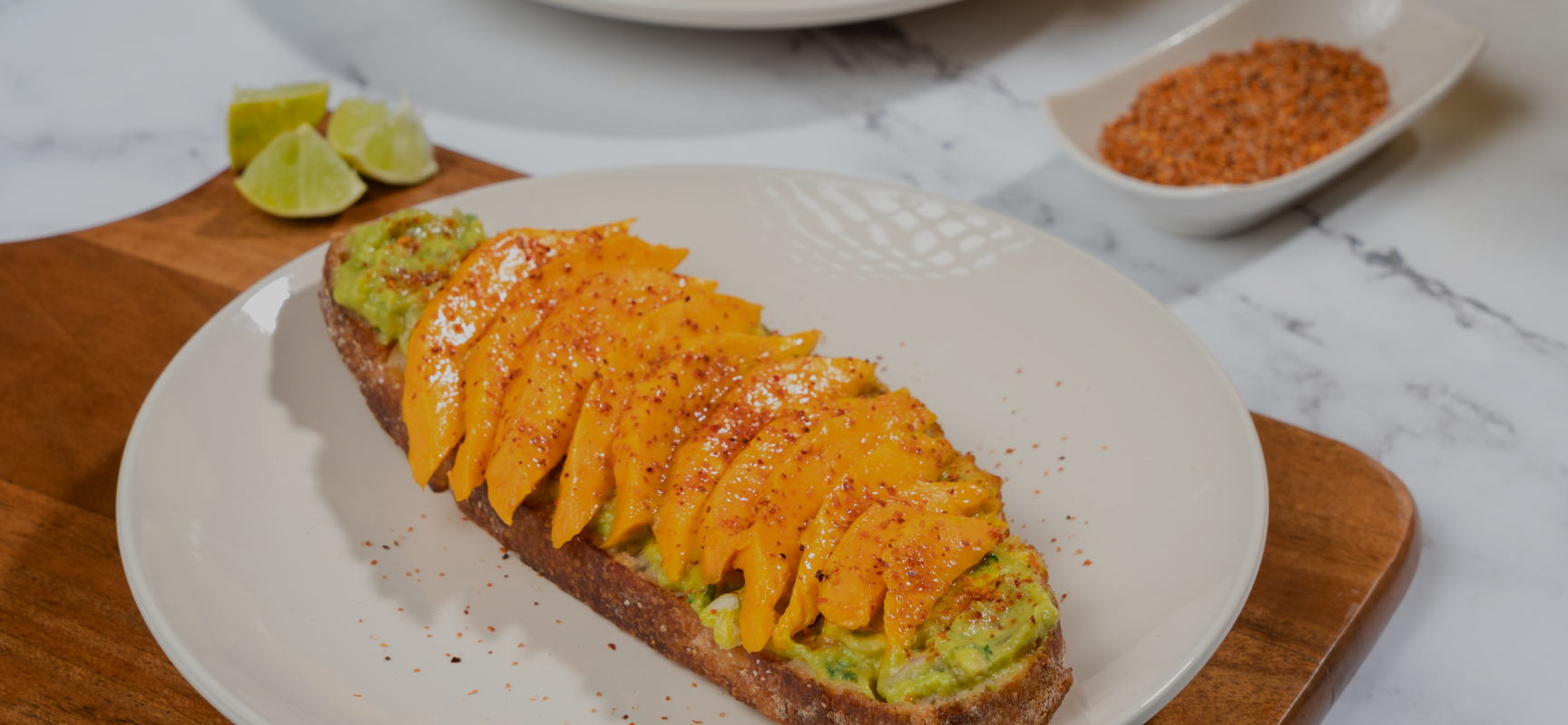 Avocado toast topped with sliced Alphonso mango, chilli seasoning, and lime wedges on a plate with a side of spices. order alphonso mangoes online