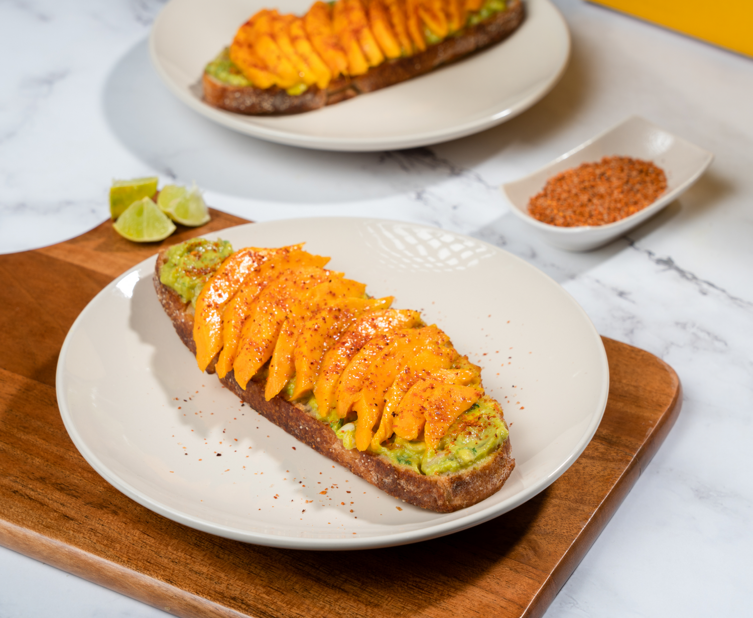 Avocado toast topped with sliced Alphonso mango, chilli seasoning, and lime wedges on a plate with a side of spices. farm fresh alphonso mango online